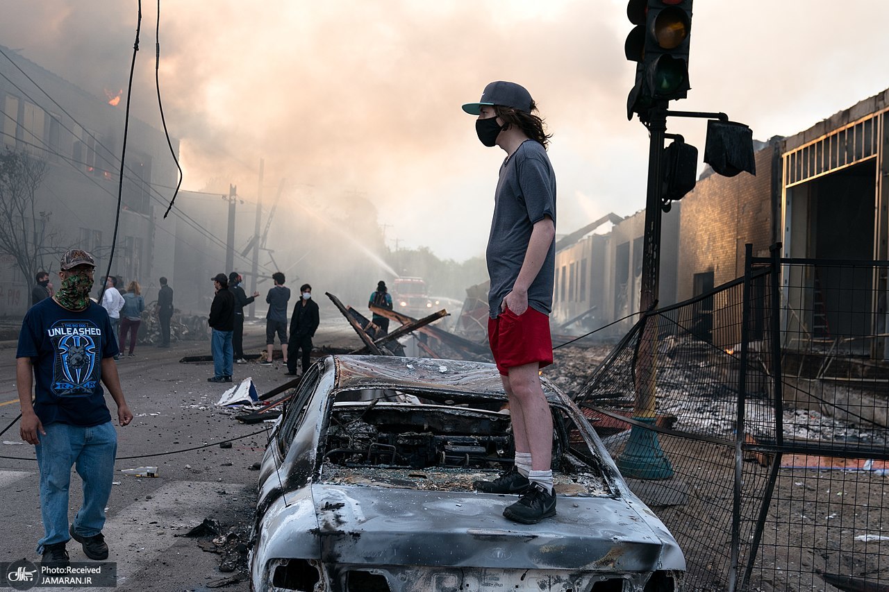 1280px-A_man_stands_on_a_burned_out_car_on_Thursday_morning_as_fires_burn_behind_him_in_the_Lake_St_area_of_Minneapolis__Minnesota_(49945886467)